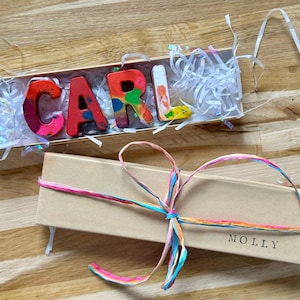 Puede incluir: Dos cajas de regalo sobre una superficie de madera. Una caja contiene letras de crayones de colores que deletrean "CARL". La otra caja está atada con una cinta multicolor y tiene el nombre "MOLLY" impreso. Los crayones son de varios colores.
