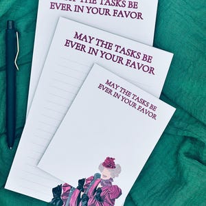 May include: Three white notepads with the text "MAY THE TASKS BE EVER IN YOUR FAVOR" in burgundy. One notepad features a pink and black illustration of a woman. A black pen rests on a green fabric background.