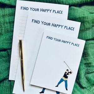 May include: Three white notepads with the text "FIND YOUR HAPPY PLACE" in teal. One notepad features a golfer illustration. A gold pen rests on the notepads. The background is a green, textured fabric.