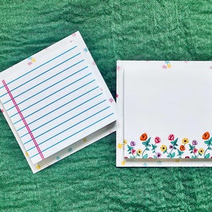 May include: Two white lined paper notepads with floral borders. The top notepad has a pink line down the center. The bottom notepad has a border of colorful flowers.