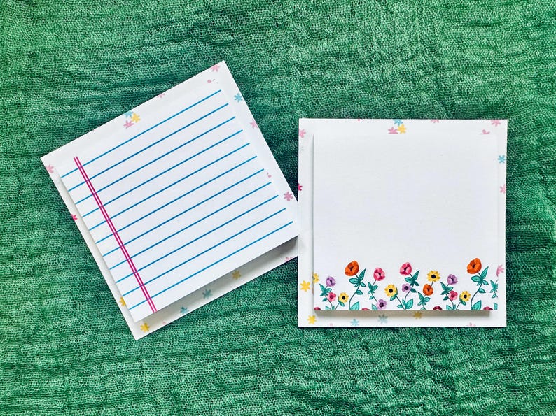 May include: Two white lined paper notepads with floral borders. The top notepad has a pink line down the center. The bottom notepad has a border of colorful flowers.