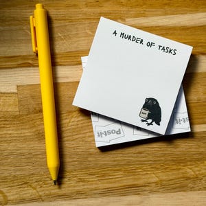 May include: A bright yellow retractable pen positioned next to a square white sticky note pad. The top sheet features the black text A MURDER OF TASKS and a small illustration of a black crow holding a mug with the word NAP printed on it.