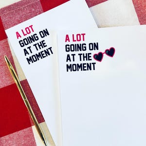 Puede incluir: Dos blocs de notas blancos con el texto "A LOT GOING ON AT THE MOMENT" en negro y rosa. Un bloc de notas tiene un par de gafas de sol en forma de corazón rosa con lentes negros.