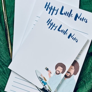 May include: Two white notepad sets with blue lined paper and the text "Happy Little Notes" printed on the front. The top notepad features a cartoon illustration of Bob Ross painting.