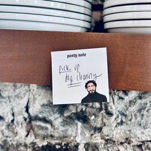 May include: A white sticky note with the text "post-it note" and "pick up dry cleaning" written in black marker. A cartoon image of a man with a beard is in the bottom right corner of the note.