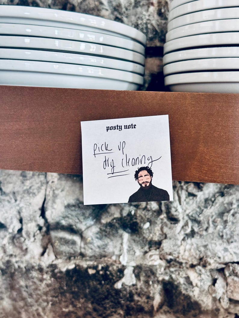 May include: A white sticky note with the text "post-it note" and "pick up dry cleaning" written in black marker. A cartoon image of a man with a beard is in the bottom right corner of the note.