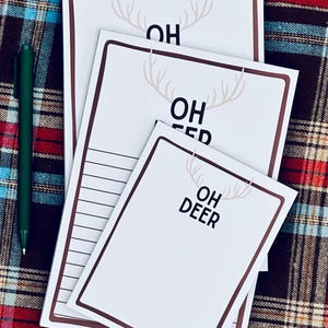 Peut inclure: Plusieurs blocs-notes blancs avec des bordures marron et le texte "OH DEER" en noir. Un motif de bois de cerf est présent. Un stylo vert est posé sur un fond à carreaux.