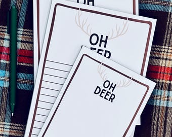 May include: A stack of white notepads with brown borders and the text "OH DEER" in black. The notepads have a deer antler graphic above the text. A green pen rests on the plaid background.