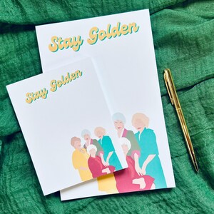 K&ouml;nnte beinhalten: Zwei wei&szlig;e Notizbl&ouml;cke mit dem Text "Stay Golden" in Gelb und Hellblau. Die Notizbl&ouml;cke zeigen eine farbenfrohe Illustration von sechs Frauen. Ein goldener Stift liegt daneben.