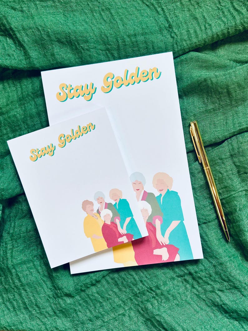 K&ouml;nnte beinhalten: Zwei wei&szlig;e Notizbl&ouml;cke mit dem Text "Stay Golden" in Gelb und Hellblau. Die Notizbl&ouml;cke zeigen eine farbenfrohe Illustration von sechs Frauen. Ein goldener Stift liegt daneben.