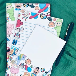 Może przedstawiać: Trzy białe kartki papieru do listów z retro motywem z lat 80. Papier ma liniowaną ramkę z kreskówkowymi obrazkami popularnych zabawek i gier z lat 80., w tym Care Bears, Cabbage Patch Kids, My Little Pony i grę Connect Four.