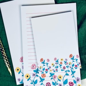 May include: Three white stationery notepads with a floral border. The border features pink, orange, yellow, and purple flowers with green leaves and stems on a white background. The top notepad is blank, the middle notepad has red lines, and the bottom notepad has a floral border.