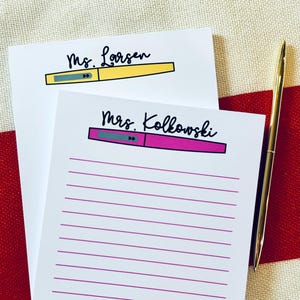 May include: Two white notepads with lined paper. The top notepad has the text "Ms. Larsen" and a yellow pen with a grey cap. The bottom notepad has the text "Mrs. Kolkowski" and a pink pen with a grey cap.