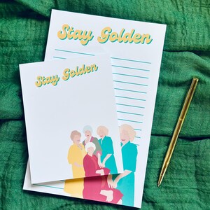 K&ouml;nnte beinhalten: Zwei wei&szlig;e Notizbl&ouml;cke mit dem Text "Stay Golden" in gelben und t&uuml;rkisfarbenen Buchstaben. Ein Notizblock hat ein liniertes Design, und der andere eine Illustration von f&uuml;nf Frauen. Ein goldener Stift liegt auf einem gr&uuml;nen Stoffhintergrund.