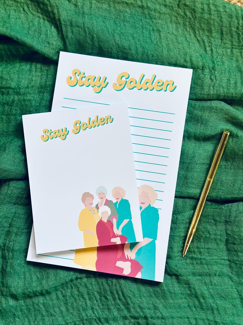 K&ouml;nnte beinhalten: Zwei wei&szlig;e Notizbl&ouml;cke mit dem Text "Stay Golden" in gelben und t&uuml;rkisfarbenen Buchstaben. Ein Notizblock hat ein liniertes Design, und der andere eine Illustration von f&uuml;nf Frauen. Ein goldener Stift liegt auf einem gr&uuml;nen Stoffhintergrund.