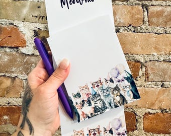 May include: A white notepad with the word "Meowmo" in dark blue script. The bottom of the notepad features a colourful illustration of cats wearing party hats, bow ties, and sunglasses. A purple pen is held next to the notepad.