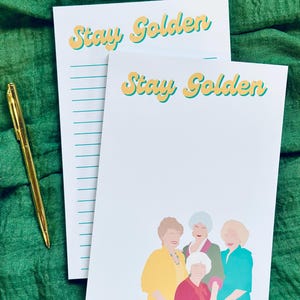 K&ouml;nnte beinhalten: Zwei wei&szlig;e Notizbl&ouml;cke mit dem Text "Stay Golden" in Gelb und T&uuml;rkis. Ein Notizblock hat linierte Seiten, der andere zeigt eine farbenfrohe Illustration von vier Frauen. Ein goldener Stift liegt auf einem gr&uuml;nen Stoff.