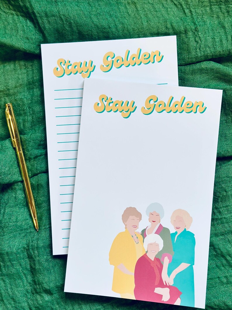 K&ouml;nnte beinhalten: Zwei wei&szlig;e Notizbl&ouml;cke mit dem Text "Stay Golden" in Gelb und T&uuml;rkis. Ein Notizblock hat linierte Seiten, der andere zeigt eine farbenfrohe Illustration von vier Frauen. Ein goldener Stift liegt auf einem gr&uuml;nen Stoff.