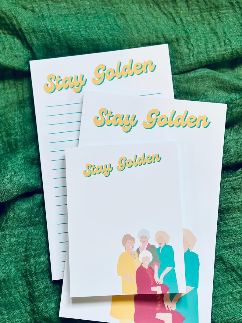 K&ouml;nnte beinhalten: Wei&szlig;e Notizbl&ouml;cke mit dem Text "Stay Golden" in gelber und t&uuml;rkisfarbener Schrift. Ein Notizblock hat linierte Seiten. Ein weiterer Notizblock zeigt eine farbenfrohe Illustration von f&uuml;nf Frauen. Die Notizbl&ouml;cke sind auf einem gr&uuml;nen Stoffhintergrund gestapelt.