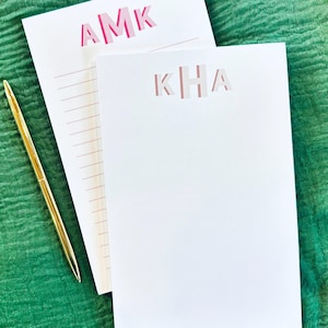 May include: Two white notepad sets with pink letters. The top notepad has the letters "AMK" and the bottom notepad has the letters "KHA". A gold pen is lying on a green surface.