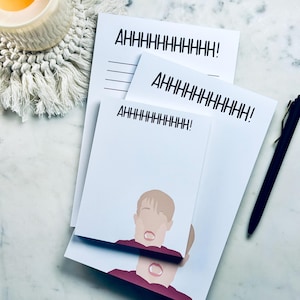 Peut inclure: Bloc-notes blancs avec le texte "AHHHHHHHHHH!" en noir. L'un des blocs-notes présente une illustration de dessin animé d'une personne avec la bouche ouverte. Un stylo noir et une bougie allumée dans un pot en verre sont également visibles.