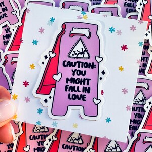 May include: A pink and red caution sign sticker with the text "CAUTION: YOU MIGHT FALL IN LOVE". The sticker is surrounded by smaller pink and red caution signs with the same text. The background is white with a pattern of small pink, blue, and yellow flowers.