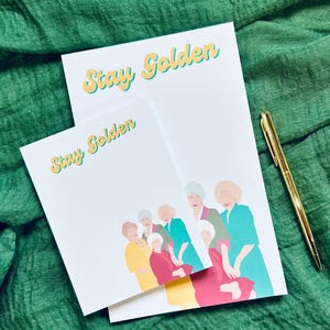 K&ouml;nnte beinhalten: Zwei wei&szlig;e Notizbl&ouml;cke mit dem Text "Stay Golden" in Gelb und Hellblau. Die Notizbl&ouml;cke zeigen eine farbenfrohe Illustration einer Frauengruppe. Ein goldener Stift liegt auf einem gr&uuml;nen Stoffhintergrund.