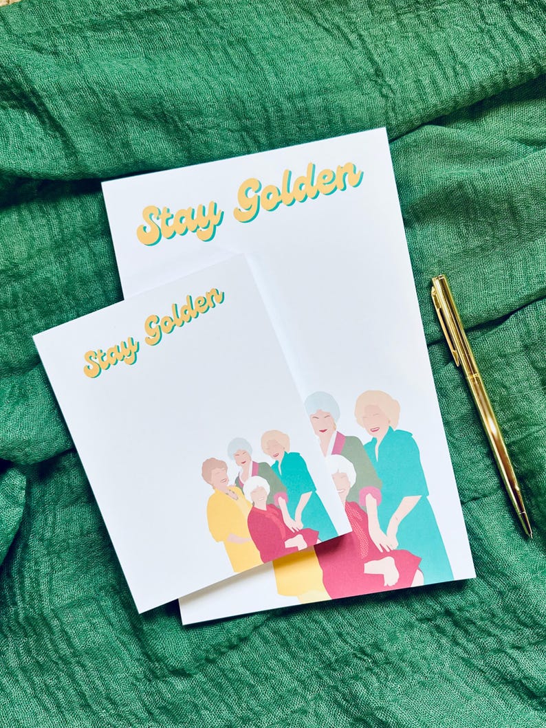K&ouml;nnte beinhalten: Zwei wei&szlig;e Notizbl&ouml;cke mit dem Text "Stay Golden" in Gelb und Hellblau. Die Notizbl&ouml;cke zeigen eine farbenfrohe Illustration einer Frauengruppe. Ein goldener Stift liegt auf einem gr&uuml;nen Stoffhintergrund.