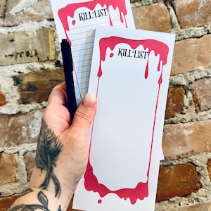 May include: White notepads with a pink dripping border and the words "KILL LIST" in black. A black pen is held with the notepads. The background is a brick wall.