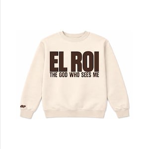 以下が含まれることがあります： クリーム色のスウェットシャツ。茶色の文字で「EL ROI THE GOD WHO SEES ME」と書かれています。クルーネックと長袖です。「10 ULTIMATE CREW OZ SWEATSHIRT」の文字が上に、「POWER OF PRAISE」の文字が下にあります。