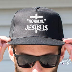 May include: Black snapback cap with the text "NORMAL ISN'T COMING BACK JESUS IS." and a cross graphic in white. The cap is being held up by a person wearing sunglasses.