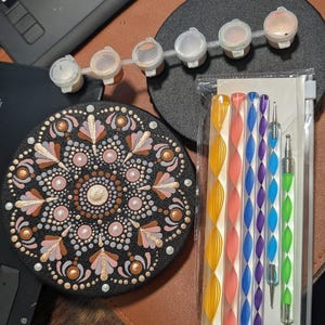 May include: A black mandala art kit with a detailed, circular design in pink, gold, and white. The kit includes paint pots, dotting tools, and a canvas. The tools are in a rainbow of colours.