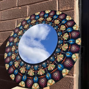 May include: Circular decorative mirror with a black frame. The frame is adorned with a colourful, dotted floral design in blue, green, orange, and pink. The mirror reflects a blue sky with clouds.