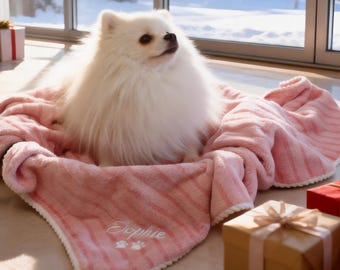 May include: A fluffy white Pomeranian dog sits in a pink striped pet bed. The bed has a white trim and the name "Sophie" embroidered on it, along with paw prints. Gift boxes are nearby.