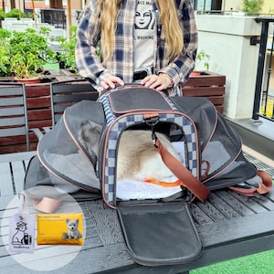 May include: A black and gray pet carrier with a checkered pattern, open to reveal a white dog resting inside. The carrier has mesh sides and a brown handle. A tag and pet waste bags are visible.