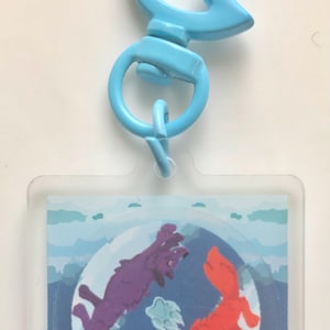 May include: A blue plastic keyring with a clear square charm featuring a red and purple wolf design. The wolves are facing each other with their mouths open, and the design is set against a blue background with white clouds.