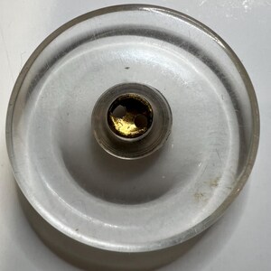 May include: Clear, circular bottle stopper with a gold-colored center. The stopper has a rounded, disc-like shape with a central, cylindrical component. The gold center features two small holes. The stopper is made of clear material.