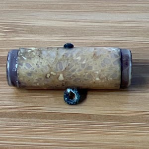 May include: A cylindrical, beige and brown bead with a dark blue accent bead. The cylindrical bead has a speckled pattern and dark brown ends. A small, dark blue bead is attached to the bottom of the cylindrical bead.