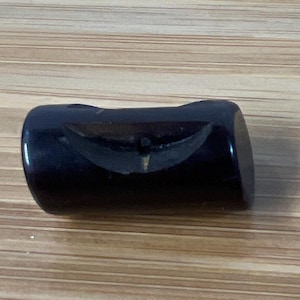 May include: A dark, rectangular object with rounded edges, possibly a bottle stopper or decorative piece. The object has a glossy, reflective surface and a subtle, curved design etched into its center. The background is a light brown, wood-grain surface.