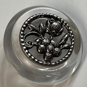 Puede incluir: Un botón redondo y transparente con un diseño floral plateado en el centro. El botón tiene una forma ligeramente abovedada y un borde decorativo alrededor del elemento floral. El diseño floral incluye uvas y hojas.