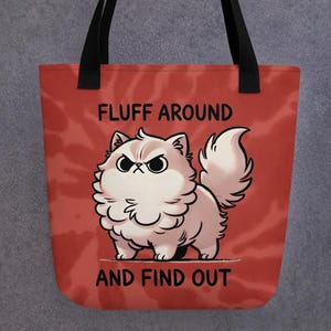 Puede incluir: Bolsa de tela de lona roja y blanca con asas negras. La bolsa presenta una ilustración de dibujos animados de un gato blanco esponjoso con una expresión gruñona. El texto "FLUFF AROUND AND FIND OUT" está impreso en negro en la bolsa.