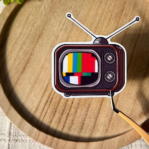 May include: A vintage-style television sticker with a colorful static screen pattern. The sticker is on a wooden surface.
