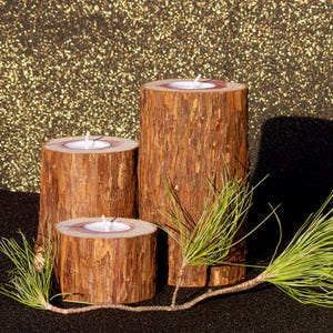 May include: Three rustic wooden candle holders of varying heights, each with a lit white candle. The holders are made from sections of tree trunks, showcasing the natural wood grain. A sprig of green pine needles rests in front of the candles, against a gold glitter background.