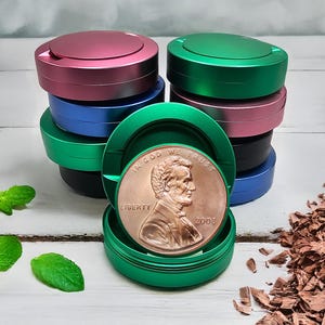 May include: A stack of colorful, round metal containers in shades of pink, green, blue, and black. One container is open, revealing a penny with the inscription "IN GOD WE TRUST" and the year 2008. The containers are on a white wooden surface.