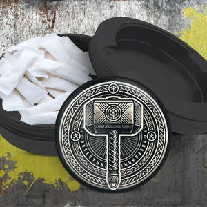 Puede incluir: Triturador negro con una tapa plateada que presenta un diseño circular con un martillo y runas. El triturador está lleno de papeles de fumar blancos.