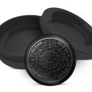 May include: A black, round container with a detailed, engraved design on the lid. The lid features a central geometric pattern surrounded by symbols and decorative elements. The container is open, revealing its interior.