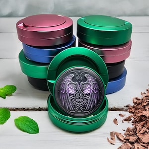 May include: A stack of colorful, round metal containers in shades of pink, green, blue, and black. One container is open, revealing a detailed, purple and black winged design. The containers are on a white surface.