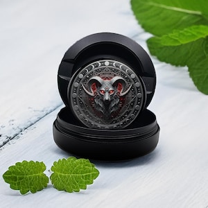 Puede incluir: Un recipiente negro con un emblema circular detallado que presenta una cabeza de carnero con ojos y cuernos rojos. El emblema está rodeado por un borde decorativo. Dos hojas verdes están en primer plano.