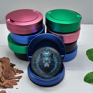 Puede incluir: Una pila de contenedores metálicos redondos y coloridos con tapa. Uno está abierto, revelando un disco negro con un diseño de lobo y detalles azules. Los contenedores son rosas, verdes, azules y negros.