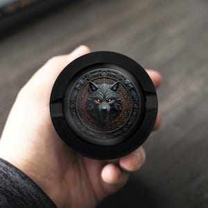 Puede incluir: Cenicero negro con un diseño detallado de cabeza de lobo. El lobo tiene ojos de color naranja brillante y está rodeado por un intrincado patrón circular. El cenicero se sostiene en una mano, con un fondo borroso.